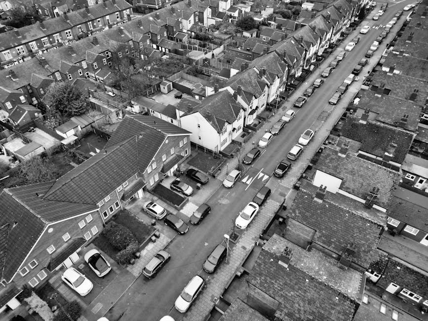 Aerial black-and-white photograph showing a row of terraced houses lining a residential street, with cars parked along both sides of the road and in driveways. The street features a narrow central lan