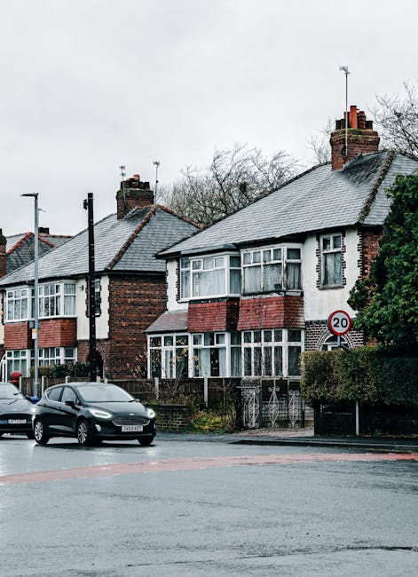 Aerial black-and-white photograph showing a row of terraced houses lining a residential street, with cars parked along both sides of the road and in driveways. The street features a narrow central lan